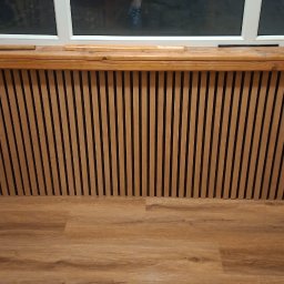 Laki - Drewniane lamele na ścianie pod oknem, z drewnianym parapetem i podłogą z paneli. Widoczny fragment narzędzi i opakowanie produktu. Wykończenie wnętrza w ciepłych barwach.