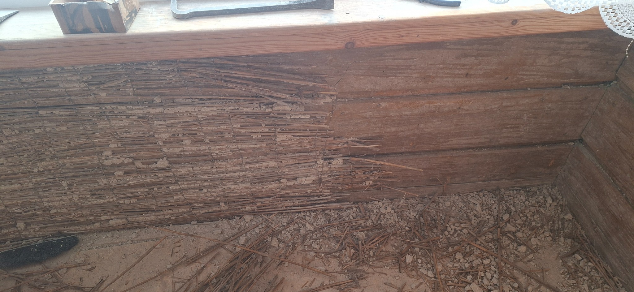 Fragment ściany z drewnianych bali z widocznym ociepleniem ze słomy i gliny, częściowo usuniętym, ukazującym konstrukcję. Narzędzia na górze kadru.