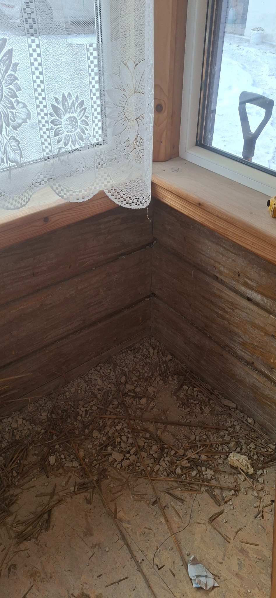 Fragment drewnianego okna z firanką i widokiem na zewnątrz, ukazujący stan podłogi przed pracami wykończeniowymi. Widoczne resztki gruzu i słomy.