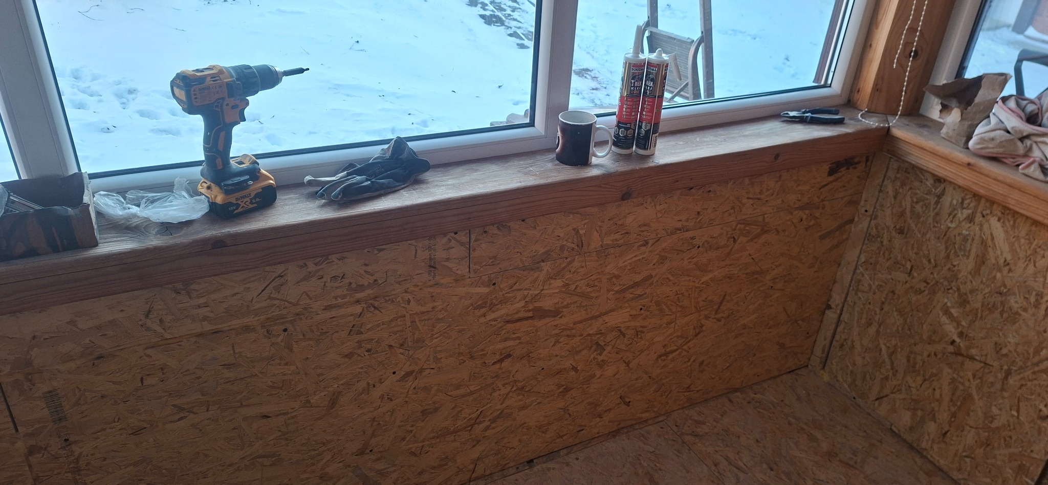 Wnętrze w trakcie wykańczania płytami OSB, widoczna wiertarka, rękawice robocze, puszki z uszczelniaczem i kubek na parapecie okna. Prace wykończeniowe.