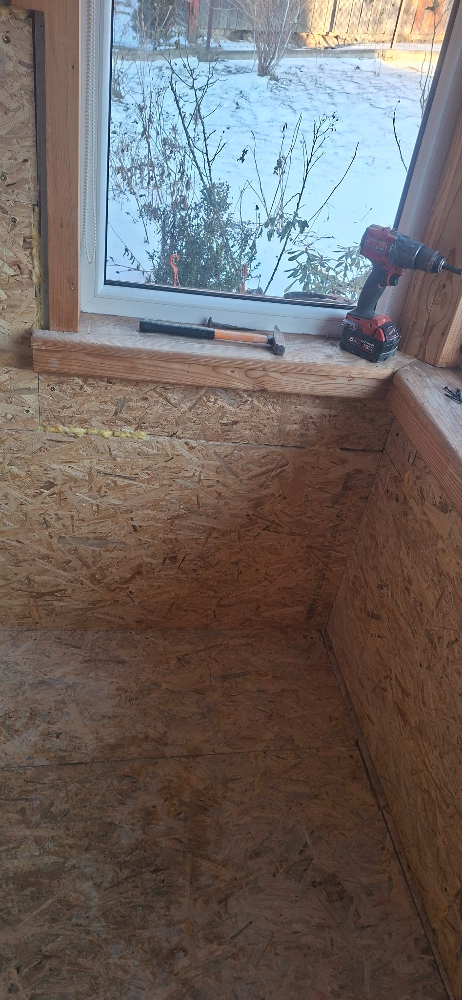 Wnętrze poddasza w trakcie wykańczania, widoczne płyty OSB, okno z widokiem na zimowy krajobraz, narzędzia: wiertarka i młotek. Prace stolarskie w surowym stanie.