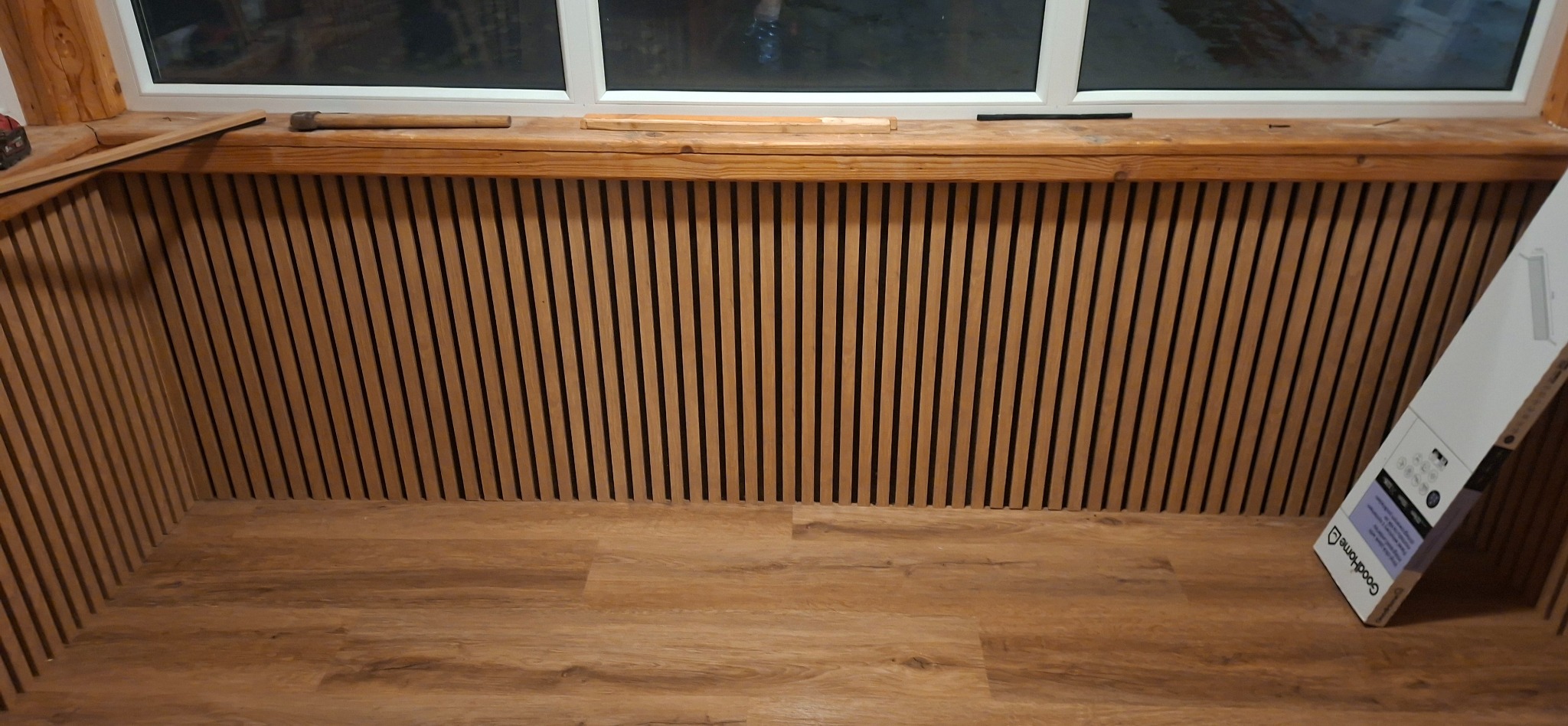 Drewniane lamele na ścianie pod oknem, z drewnianym parapetem i podłogą z paneli. Widoczny fragment narzędzi i opakowanie produktu. Wykończenie wnętrza w ciepłych barwach.