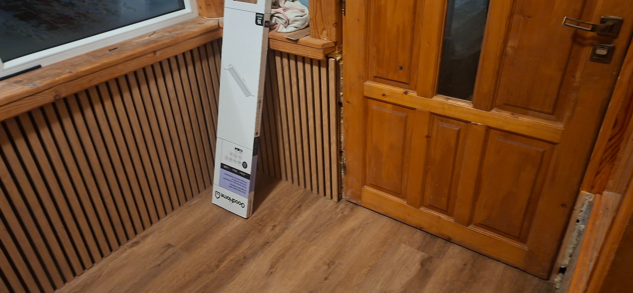 Montaż paneli ściennych z drewnianymi lamelami, widoczna paczka paneli GoodHome, drewniana podłoga i drzwi. Detal z prac wykończeniowych.