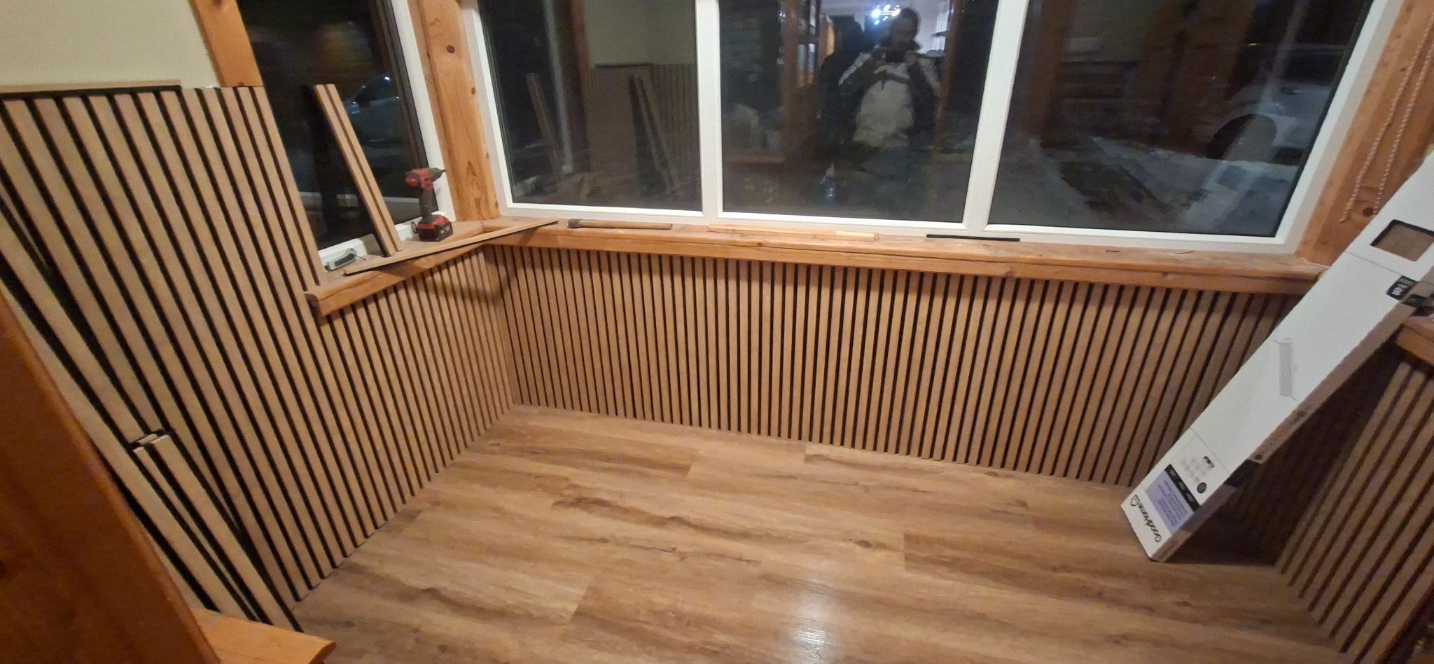 Montaż paneli ściennych z drewna w pomieszczeniu z oknami. Widoczne narzędzia i opakowanie paneli. Podłoga wyłożona panelami imitującymi drewno.