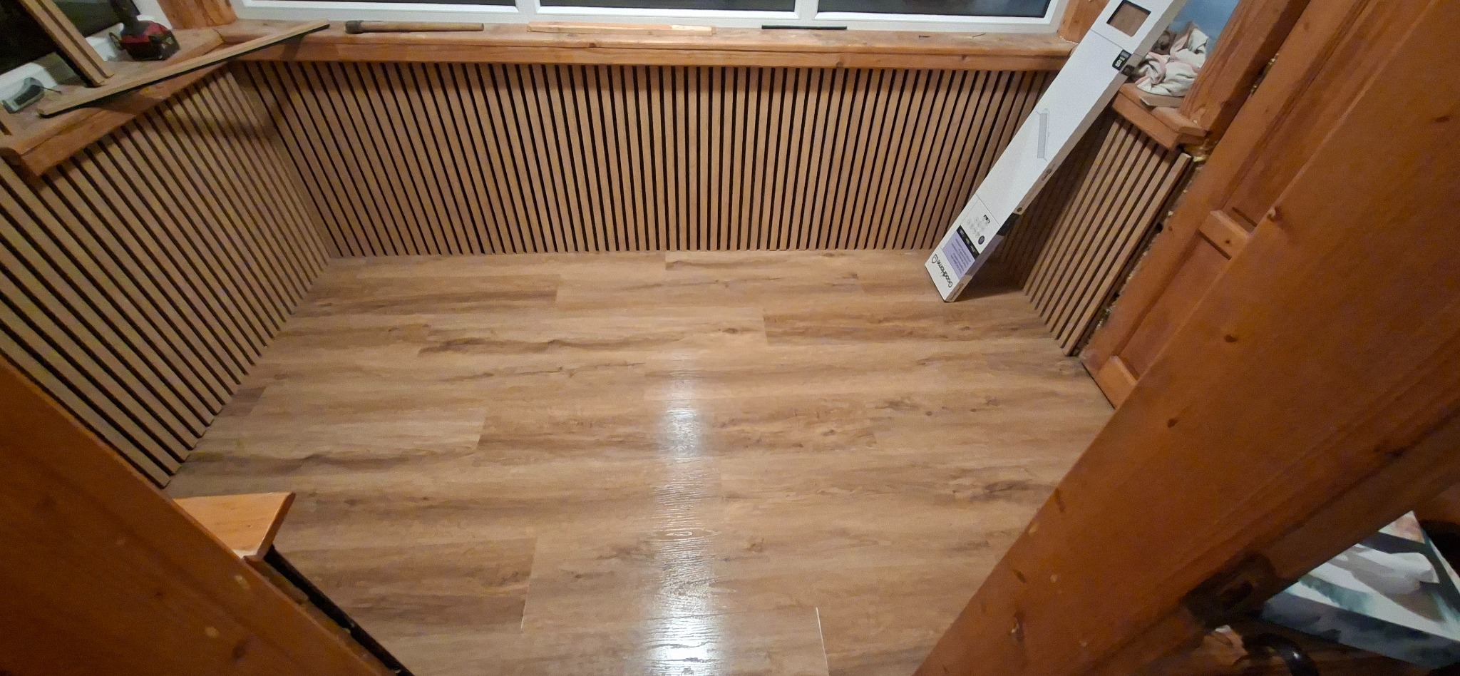 Wykończone wnętrze z panelami ściennymi i podłogą imitującą drewno. Widoczny fragment schodów i opakowanie paneli. Ciepłe oświetlenie.