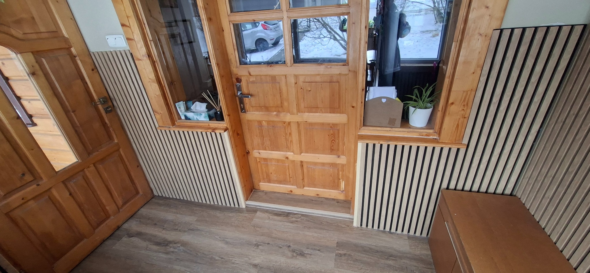 Drewniane panele ścienne z pionowymi listwami w przedpokoju, widoczne drzwi wejściowe i fragment podłogi. Dekoracyjne wykończenie wnętrza z elementami roślinnymi.