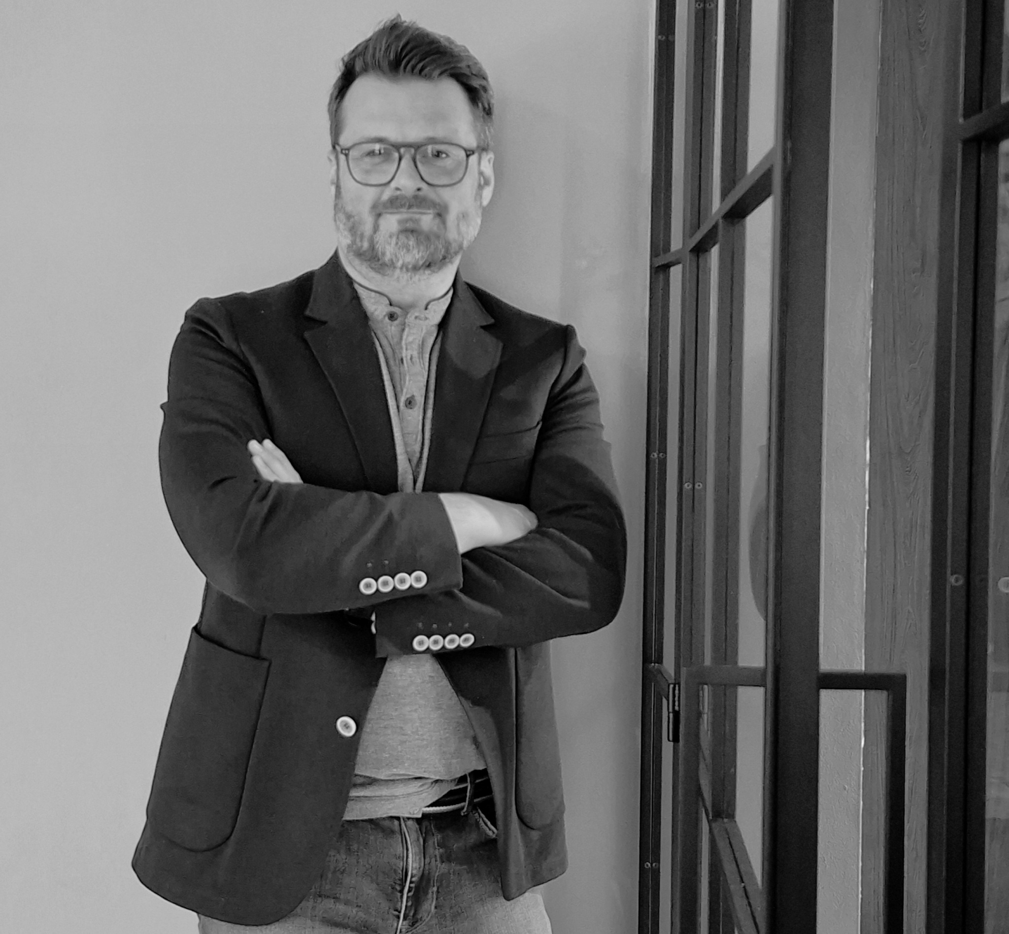 Czarno-białe zdjęcie architekta wnętrz w okularach, stojącego na tle ściany i przeszklonych drzwi, z założonymi rękami, prezentującego profesjonalny wizerunek.