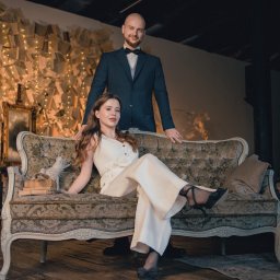 Michał Janicki - Elegancka para: kobieta w bieli siedzi na zdobionej sofie, mężczyzna w garniturze stoi za nią. W tle ściana z książek i światełek, tworząca unikalną scenografię.