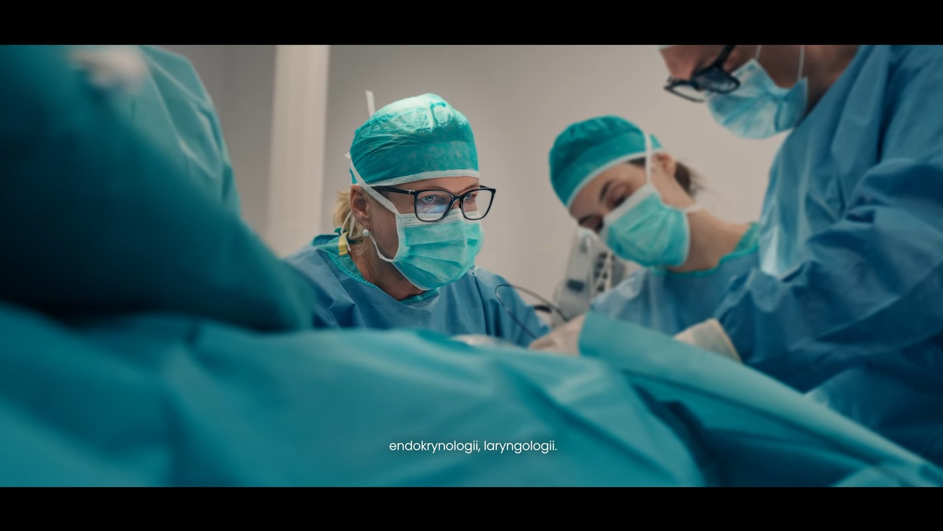 Kadr z filmu: zespół chirurgów w trakcie operacji tarczycy, w zielonych strojach i maskach, skupiony na zabiegu. Oświetlenie studyjne, precyzyjne ujęcie.