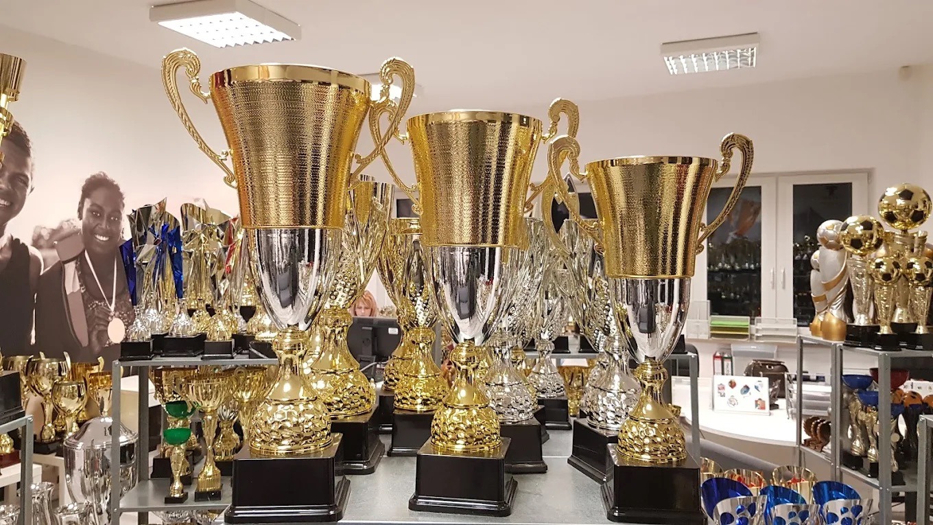 Różne puchary sportowe w kolorze złotym i srebrnym, ustawione na półkach w sklepie. W tle widoczna osoba i plakat z wizerunkiem sportowców.