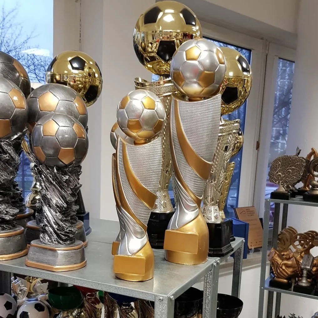Kolekcja błyszczących, srebrno-złotych trofeów piłkarskich o różnych kształtach i rozmiarach, ustawionych na metalowych półkach w jasnym pomieszczeniu.