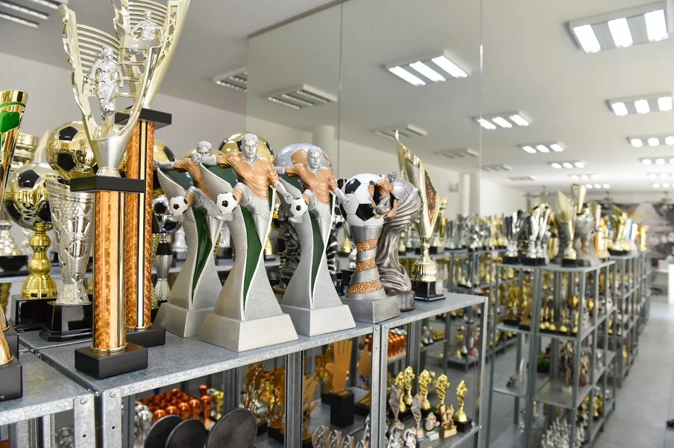 Rzędy półek z różnorodnymi trofeami sportowymi, głównie piłkarskimi, w jasnym wnętrzu sklepu. Widoczne puchary, statuetki i medale.
