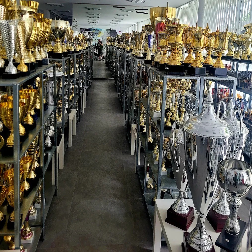 Sala pełna trofeów: złote i srebrne puchary na metalowych regałach, tworzące aleję sukcesu. Osoba w tle dodaje skali.