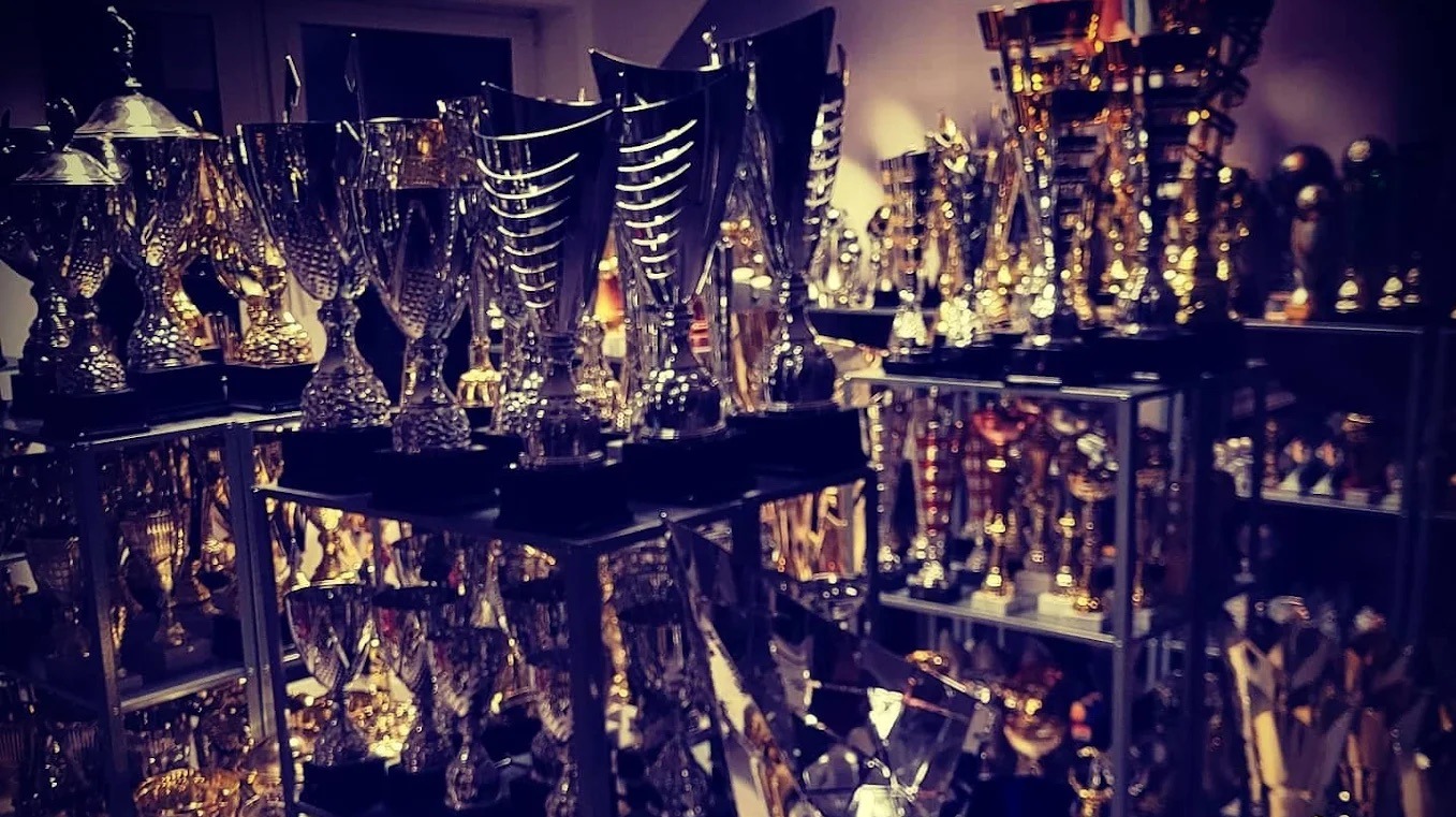 Kolekcja trofeów i pucharów na półkach, różne kształty i rozmiary, ciemne oświetlenie, odblaski. Nagrody sportowe i okolicznościowe.