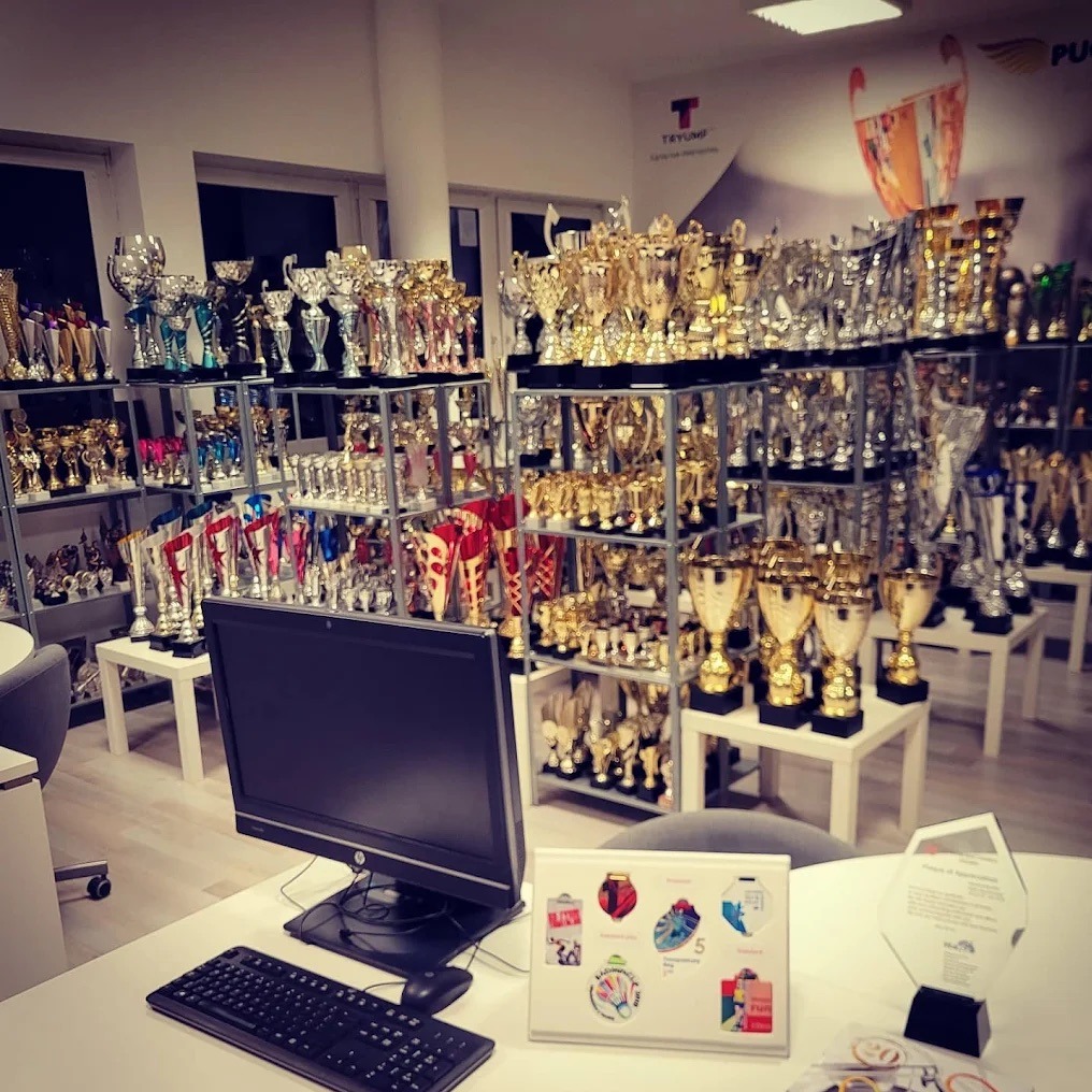 Wnętrze sklepu z trofeami: półki uginają się od złotych i srebrnych pucharów. Na pierwszym planie biurko z komputerem i ekspozycją wzorów. Stonowana kolorystyka.