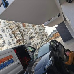 Mobilna Wulkanizacja Włocławek Serwis Mobilny - Mobilna wulkanizacja: mężczyzna w trakcie wymiany opony przy czarnym samochodzie, obok biały bus z logo firmy na tle bloku mieszkalnego.