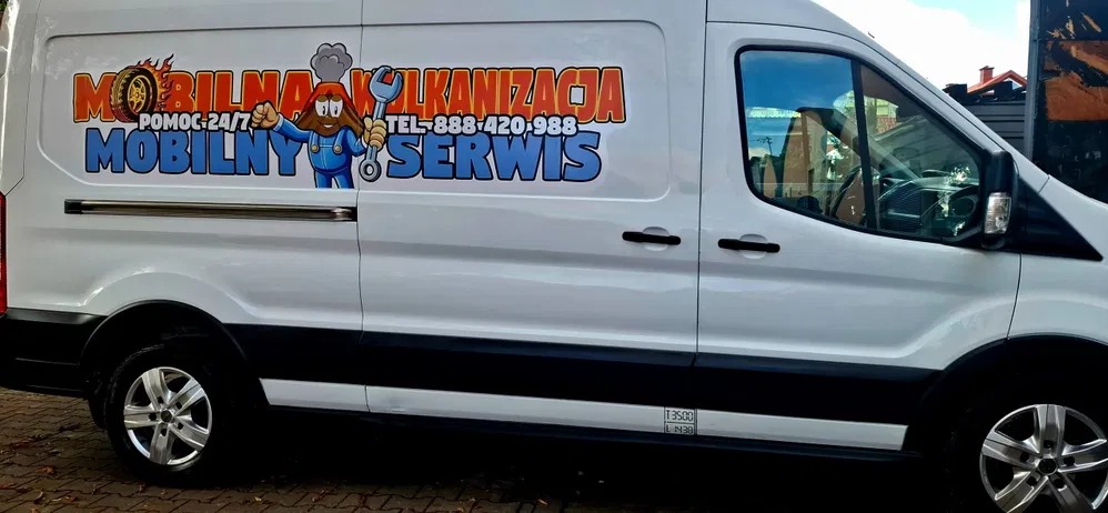Biały samochód dostawczy z kolorową grafiką 'Mobilna Wulkanizacja' i numerem telefonu, zaparkowany na zewnątrz. Widoczny bok pojazdu z felgami i odbiciem w oknie.