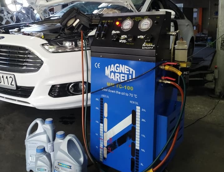 Wymiana oleju w automatycznej skrzyni biegów Ford Fusion. Urządzenie Magneti Marelli, butelki oleju Ravenol ATF. Widoczny otwarty silnik.