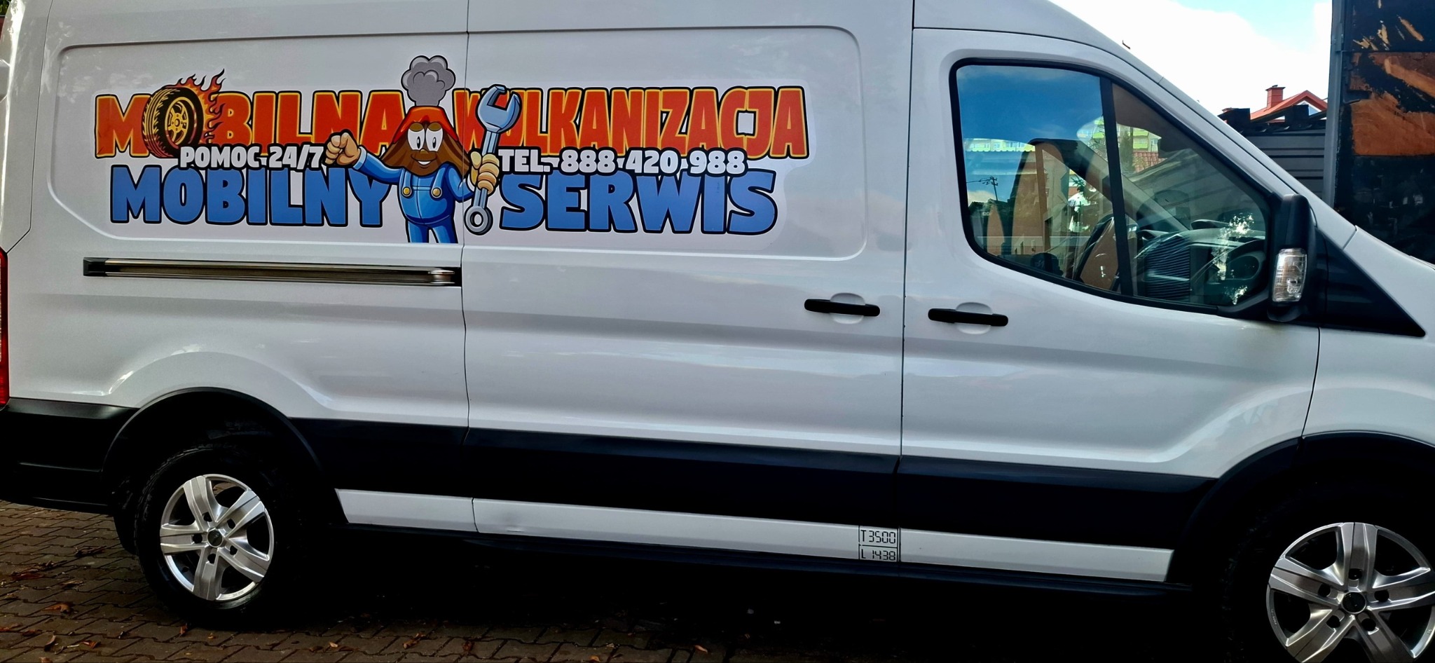 Biały bus z kolorową grafiką 'Mobilna Wulkanizacja' i wulkanem z kluczem francuskim. Usługa mobilna 24/7. Widok z boku, felgi aluminiowe.