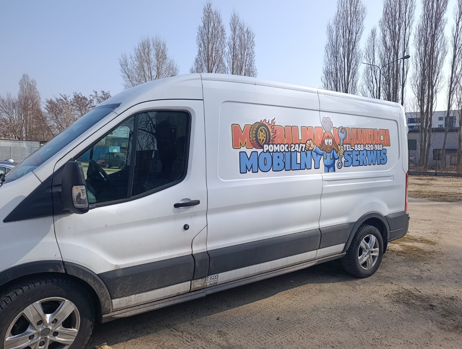 Biały bus z kolorową grafiką 'Mobilna Wulkanizacja' i numerem telefonu, zaparkowany na placu. Usługa mobilnego serwisu opon 24/7.