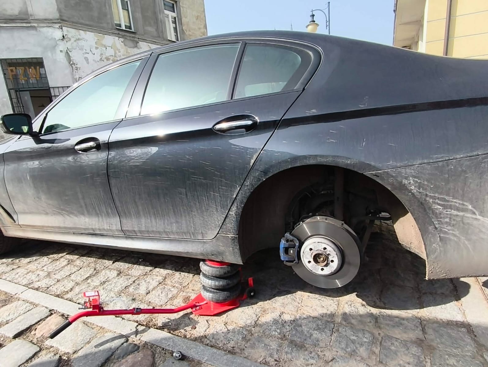 Czarny samochód z podnośnikiem pneumatycznym przy kole, na brukowanej ulicy. Widoczny fragment budynku w tle. Brak opony, tarcza hamulcowa na widoku.