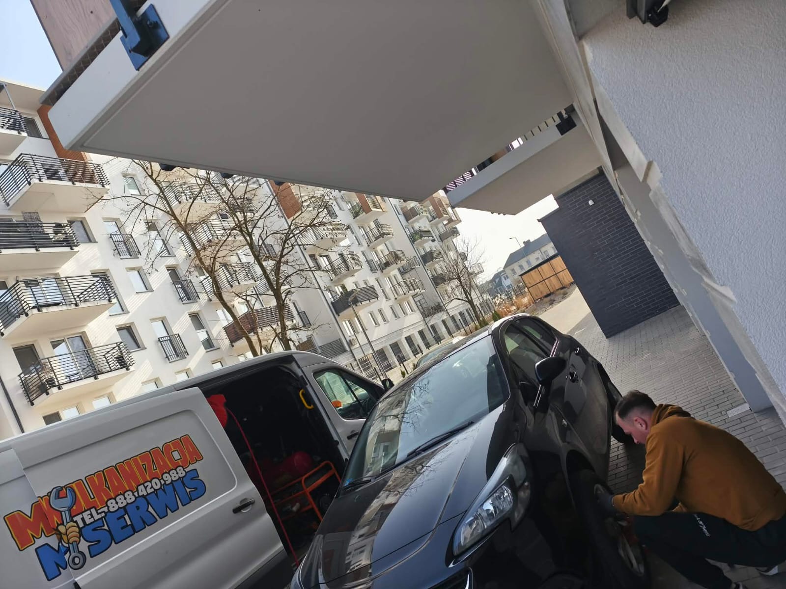 Mobilna wulkanizacja: mężczyzna w trakcie wymiany opony przy czarnym samochodzie, obok biały bus z logo firmy na tle bloku mieszkalnego.