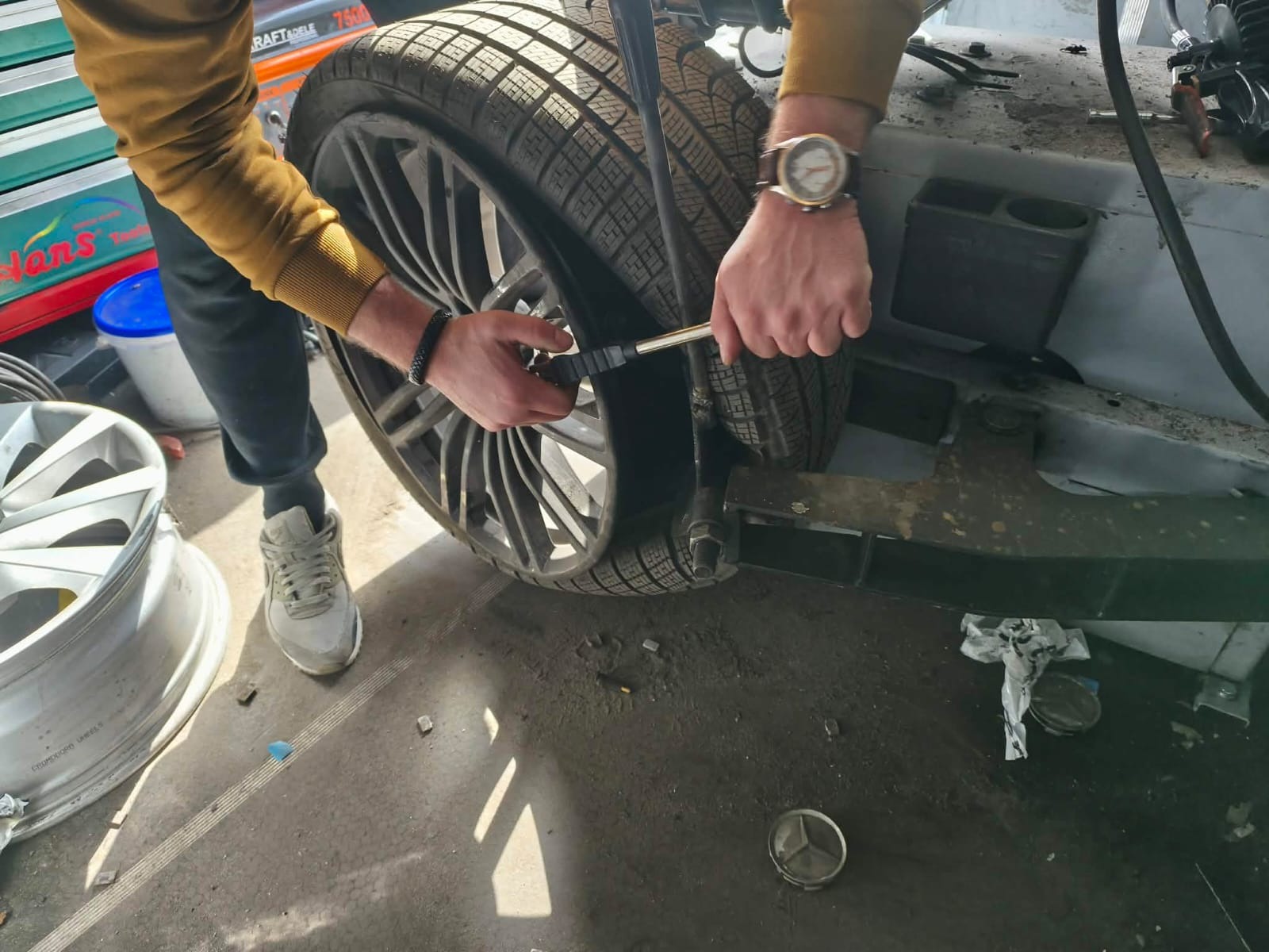 Mechanik sprawdza ciśnienie w oponie zimowej na feldze aluminiowej. Widoczny fragment podwozia samochodu i narzędzia. Mężczyzna w zegarku i sportowych butach.