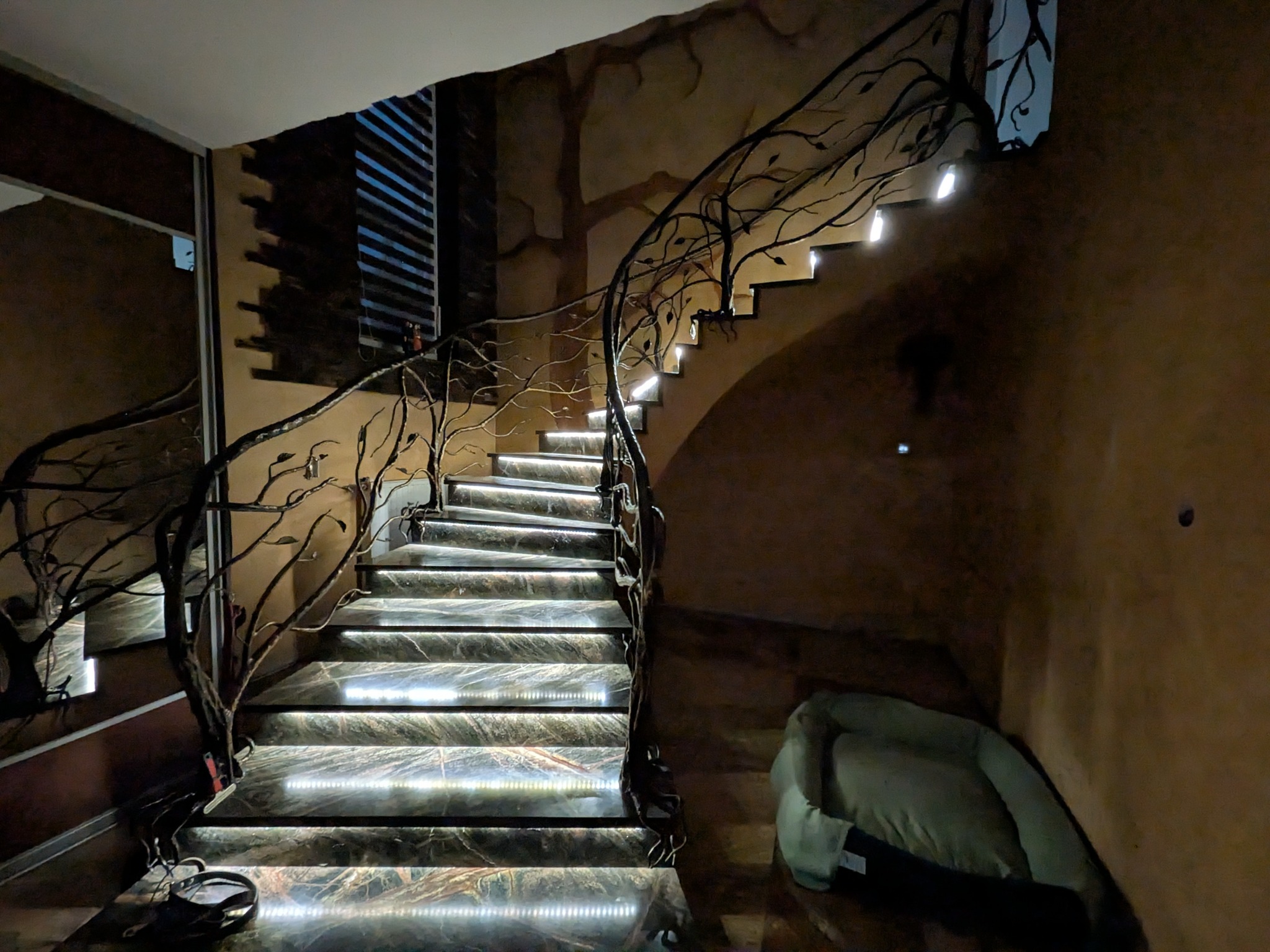 Schody z podświetleniem LED i ozdobną balustradą z motywem drzewa, prowadzące w górę. Widoczne elementy dekoracyjne na ścianie i poduszka na podłodze.