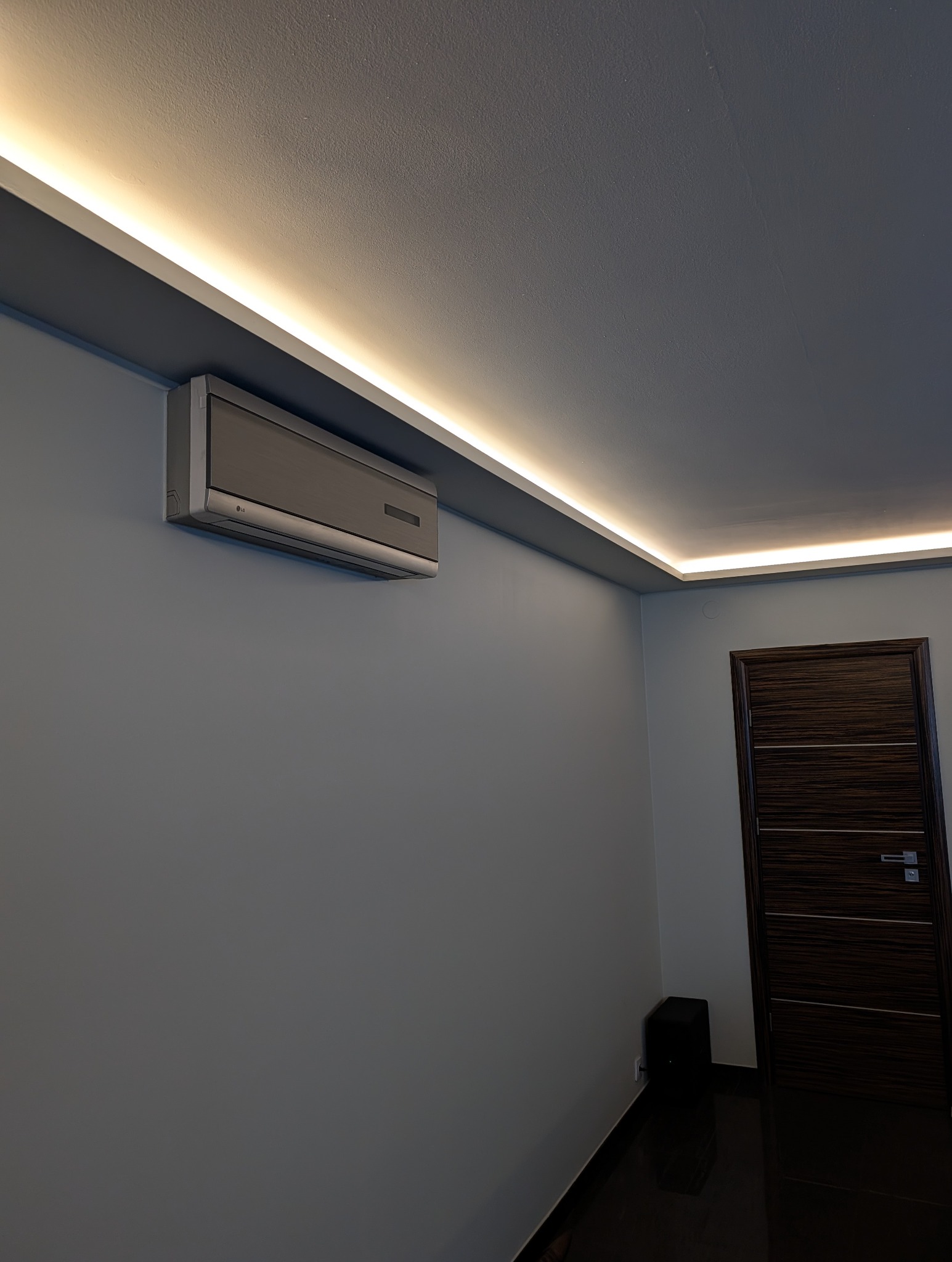 Szary klimatyzator LG na ścianie pod sufitem z oświetleniem LED. Ciemne drzwi po prawej stronie. Minimalistyczny design wnętrza.