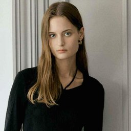 Olga Jankowska - Portret kobiety o jasnych włosach i niebieskich oczach, ubrana w czarną bluzkę, pozująca na tle jasnych drzwi. Subtelny makijaż i złote kolczyki.