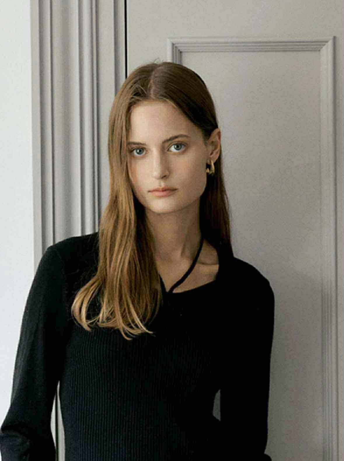 Portret kobiety o jasnych włosach i niebieskich oczach, ubrana w czarną bluzkę, pozująca na tle jasnych drzwi. Subtelny makijaż i złote kolczyki.