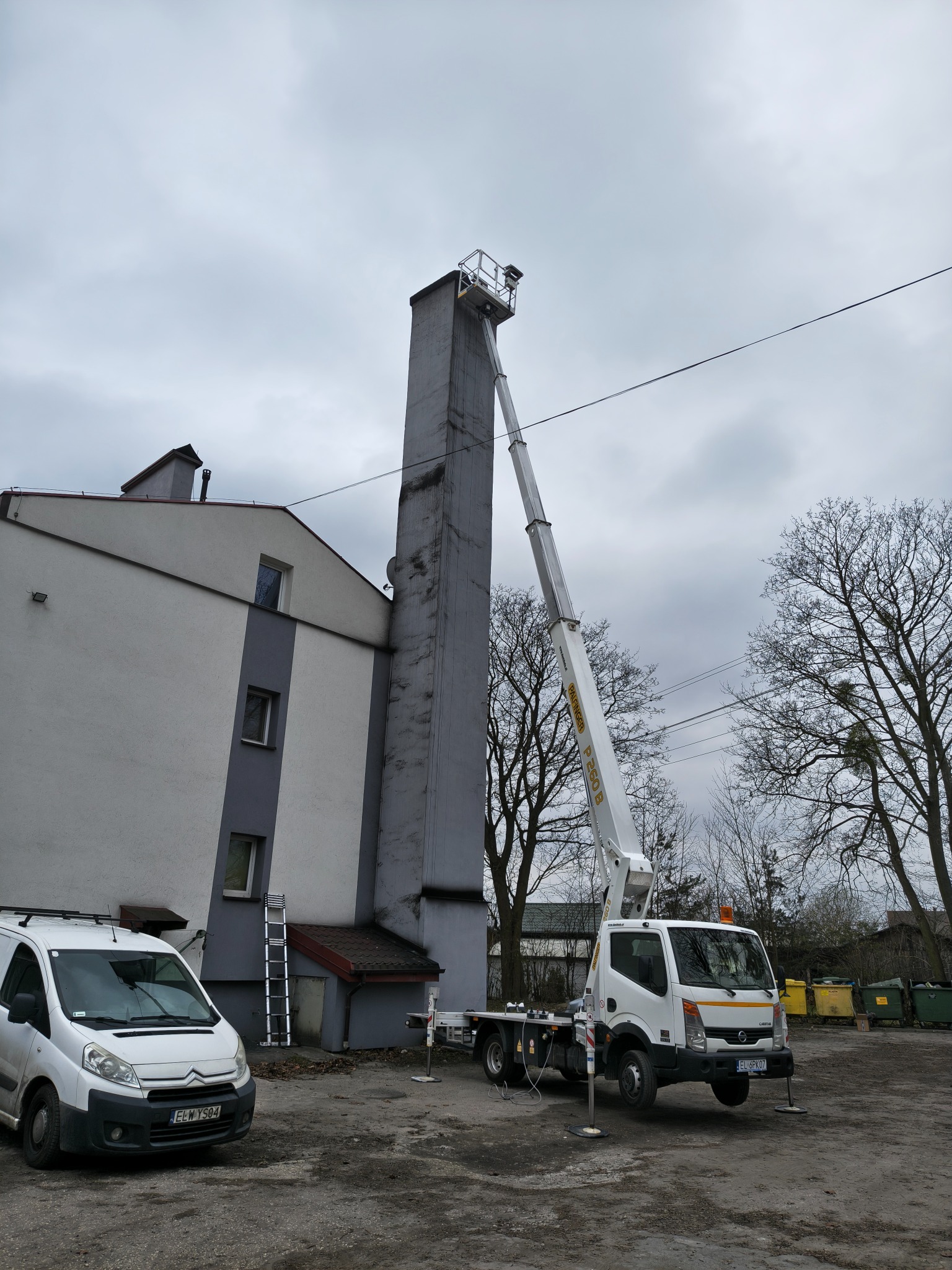 Biały podnośnik koszowy na tle budynku z kominem, wsparty na podporach. Obok zaparkowany biały bus i drabina. Pochmurne niebo.