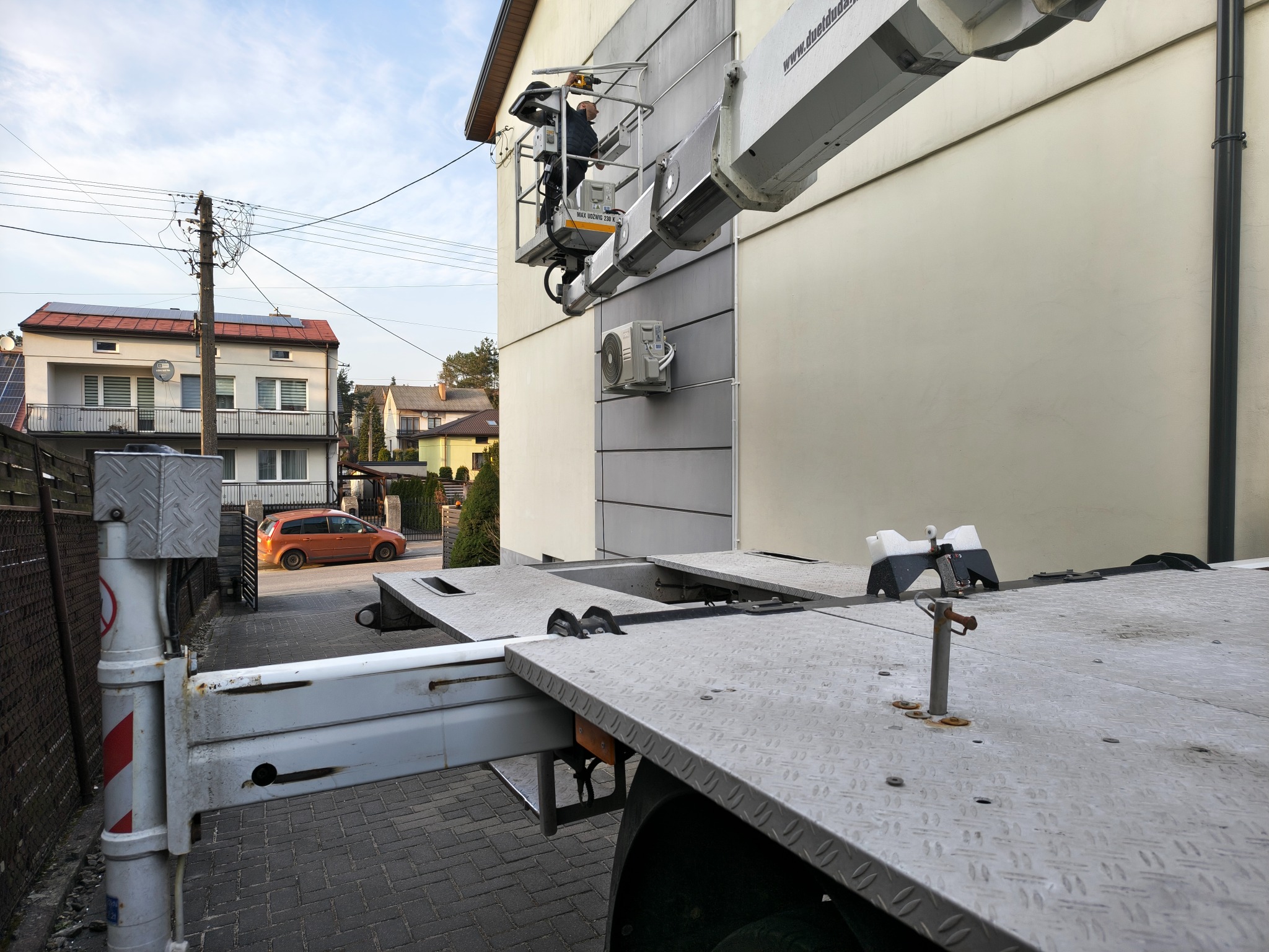 Pracownik w koszu podnośnika wykonuje prace na elewacji budynku z klimatyzacją. Widoczny fragment podwozia podnośnika i zabudowa domów w tle.