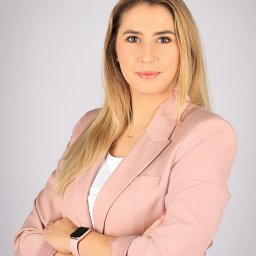 Freedom Lublin - Portret kobiety w pudrowym różu, skrzyżowane ręce, zegarek na nadgarstku, delikatny makijaż, blond włosy, subtelna biżuteria. Profesjonalny wizerunek agenta nieruchomości.