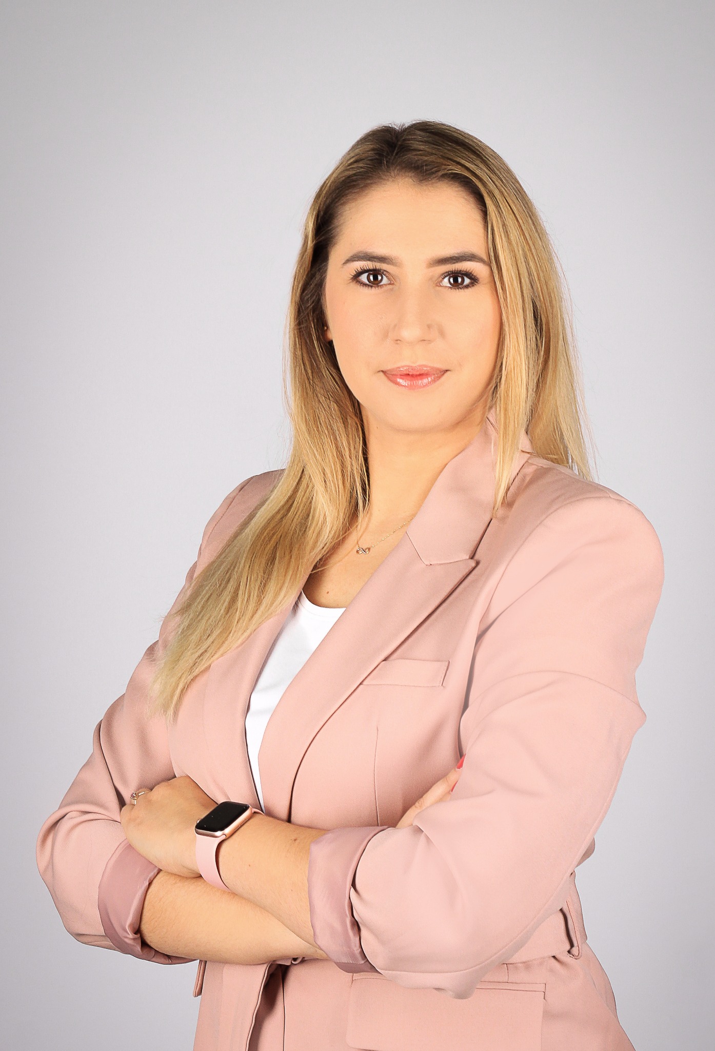 Portret kobiety w pudrowym różu, skrzyżowane ręce, zegarek na nadgarstku, delikatny makijaż, blond włosy, subtelna biżuteria. Profesjonalny wizerunek agenta nieruchomości.