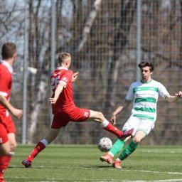 Maciej Stanowski - Dynamiczne ujęcie z meczu piłki nożnej: zawodnik w czerwonym stroju kopie piłkę w kierunku przeciwnika w zielono-białym stroju na tle zielonej murawy i siatki.