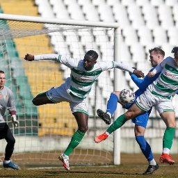 Maciej Stanowski - Mecz piłki nożnej: zawodnik w białej koszulce z zielonymi pasami w dynamicznym wyskoku, walczący o piłkę z przeciwnikiem na tle bramki i trybun.