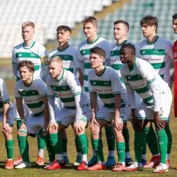 Maciej Stanowski - Drużyna piłkarska w białych koszulkach z zielonymi pasami pozuje na boisku. Widoczny bramkarz w czerwonym stroju. Ujęcie z dołu, w tle trybuny.