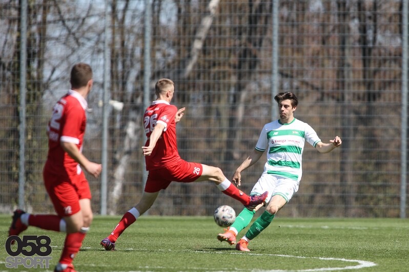 Dynamiczne ujęcie z meczu piłki nożnej: zawodnik w czerwonym stroju kopie piłkę w kierunku przeciwnika w zielono-białym stroju na tle zielonej murawy i siatki.