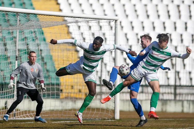 Mecz piłki nożnej: zawodnik w białej koszulce z zielonymi pasami w dynamicznym wyskoku, walczący o piłkę z przeciwnikiem na tle bramki i trybun.