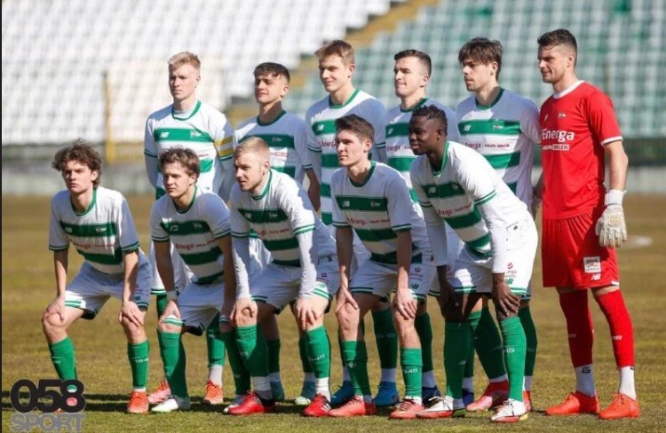 Drużyna piłkarska w białych koszulkach z zielonymi pasami pozuje na boisku. Widoczny bramkarz w czerwonym stroju. Ujęcie z dołu, w tle trybuny.