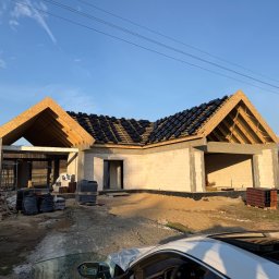 Andach Andrzej Niebieszczanski - Surowy stan domu z białego pustaka, z częściowo położonym dachem z czarnej dachówki. Widoczne drewniane elementy konstrukcji dachu i materiały budowlane na placu budowy.