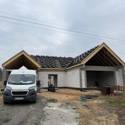 Andach Andrzej Niebieszczanski - Budowa domu jednorodzinnego w trakcie realizacji: ściany z betonu komórkowego, więźba dachowa z częściowo ułożoną dachówką. Na pierwszym planie biały bus.