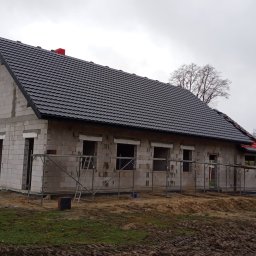 Andach Andrzej Niebieszczanski - Surowy dom z szarej cegły z czarnym dachem z dachówki, otoczony rusztowaniem. Widoczne niezabudowane otwory okienne i drzwiowe. W tle drzewa i pochmurne niebo.