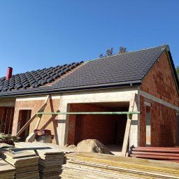 Andach Andrzej Niebieszczanski - Niedokończony dom z czerwonej cegły z częściowo ułożonym dachem z czarnej dachówki i czerwonym kominem. Widoczne materiały budowlane i drewniane podpory.