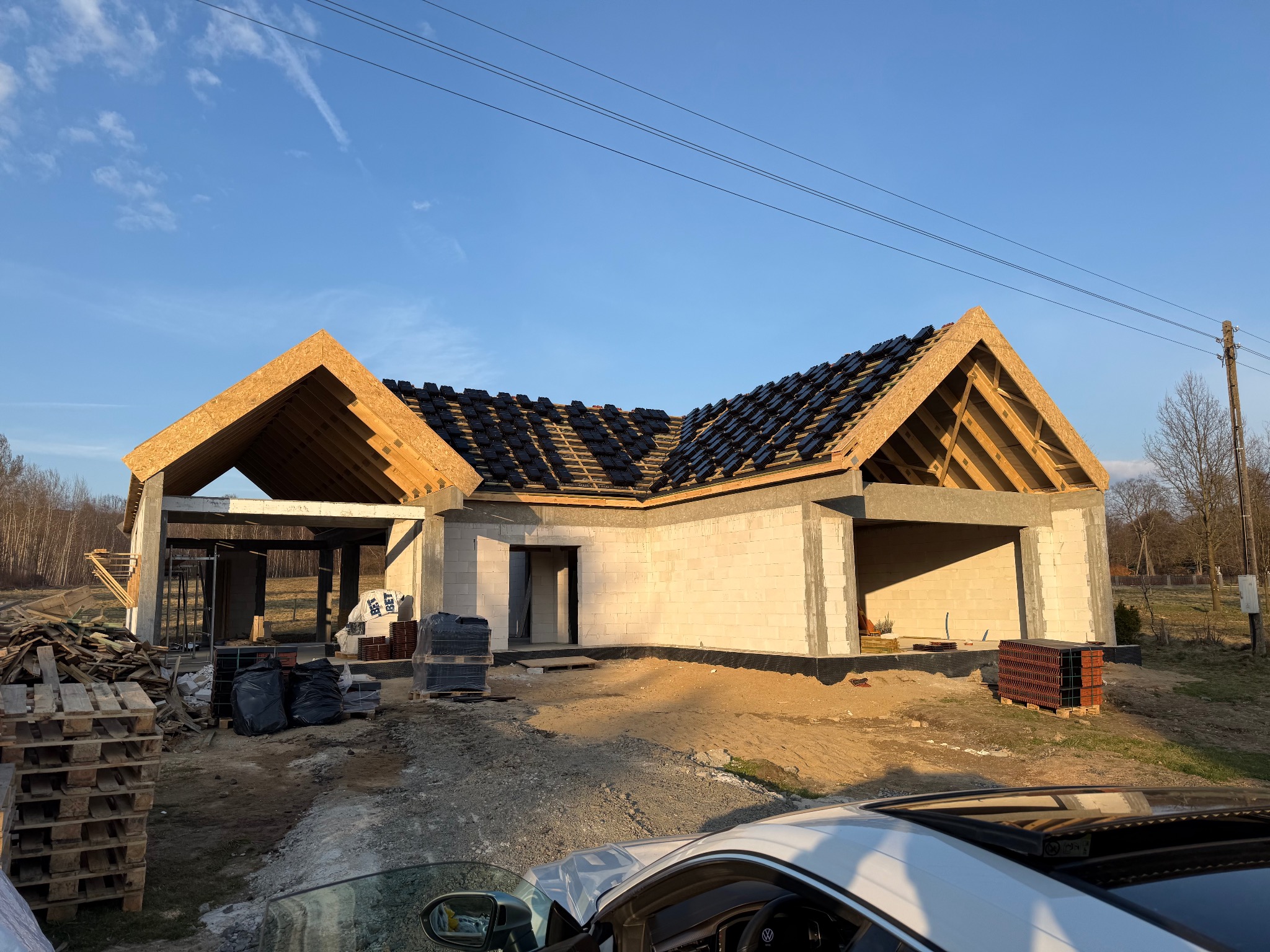 Surowy stan domu z białego pustaka, z częściowo położonym dachem z czarnej dachówki. Widoczne drewniane elementy konstrukcji dachu i materiały budowlane na placu budowy.