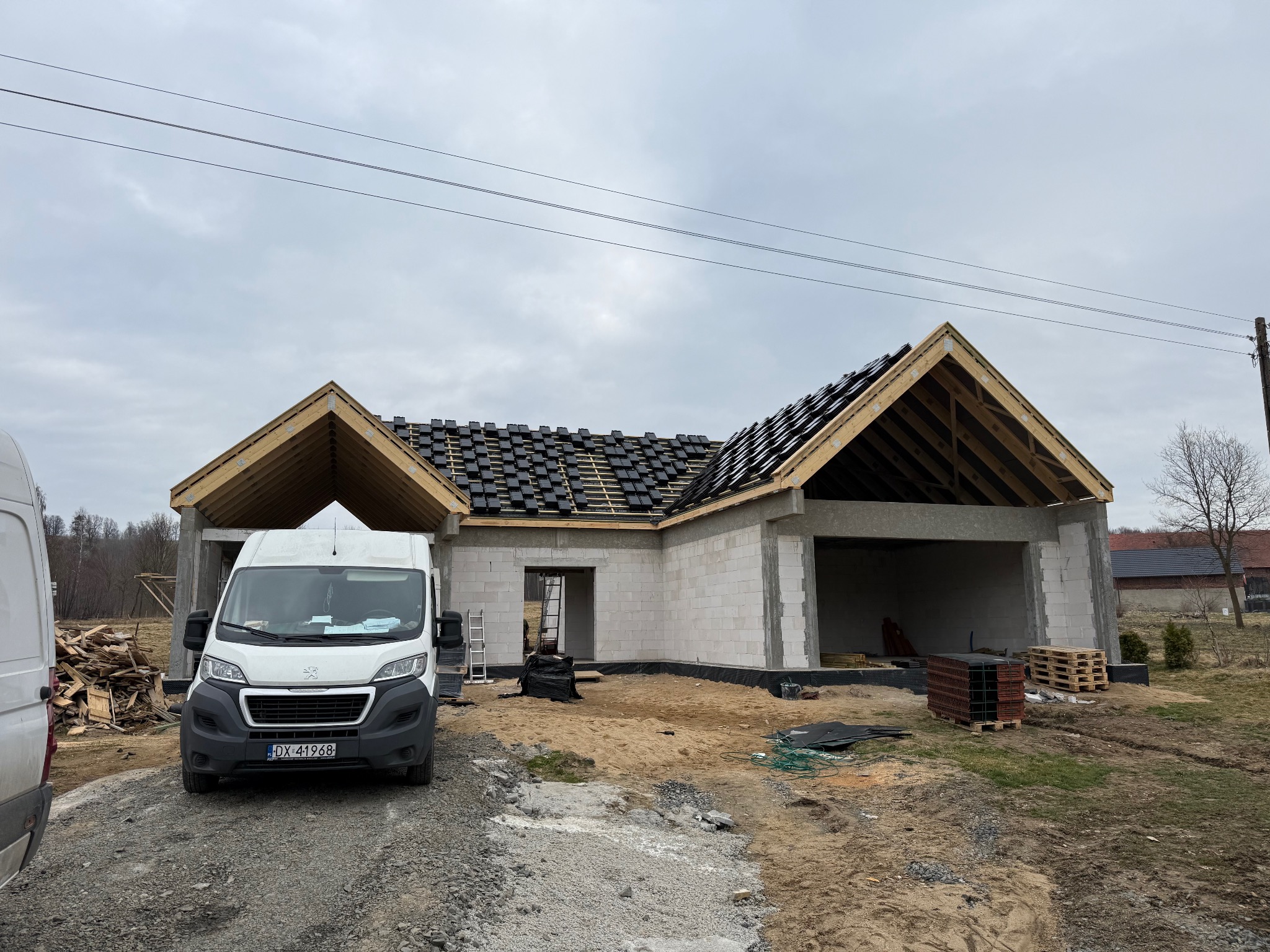 Budowa domu jednorodzinnego w trakcie realizacji: ściany z betonu komórkowego, więźba dachowa z częściowo ułożoną dachówką. Na pierwszym planie biały bus.