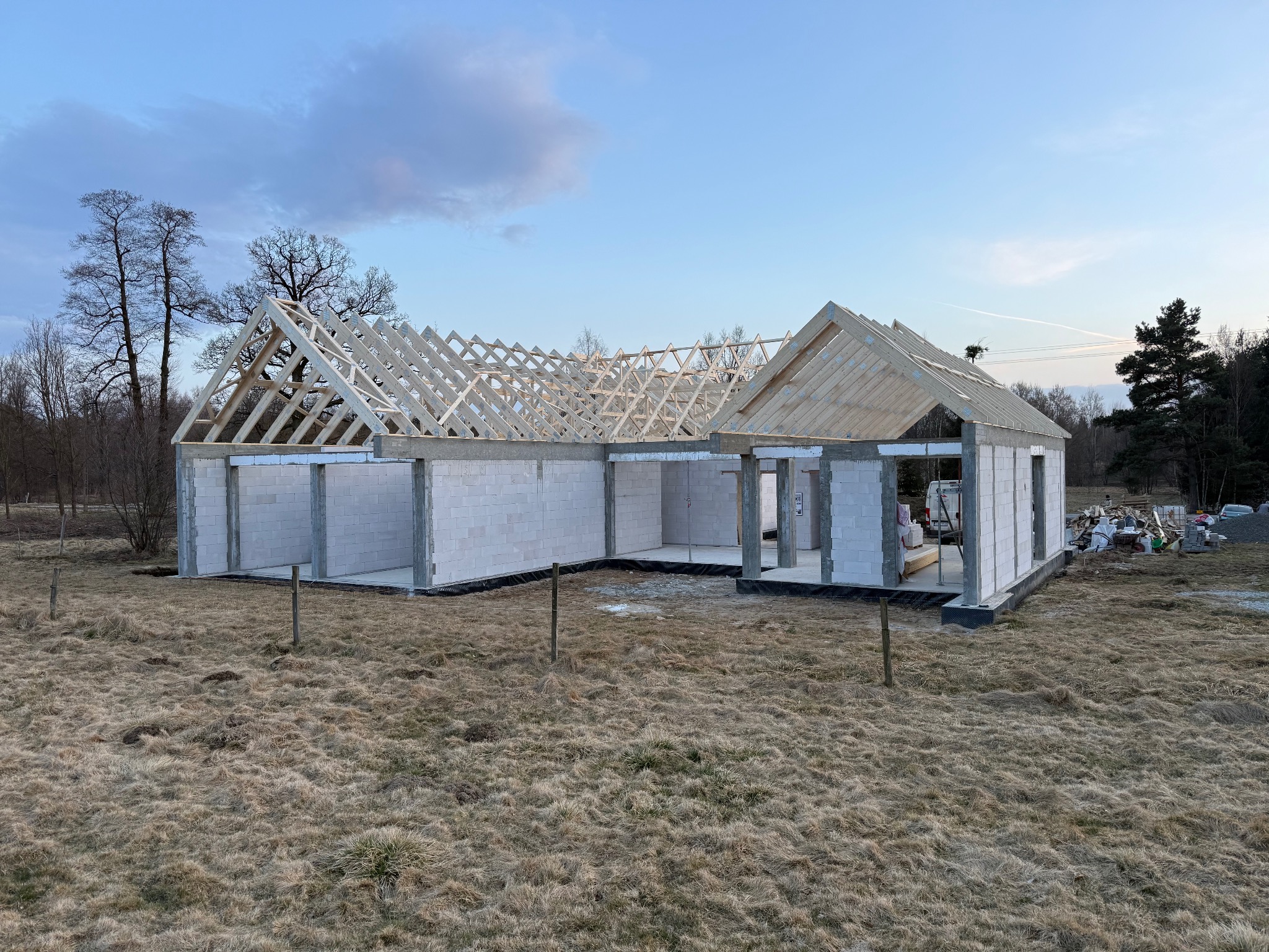 Surowy stan domu z białych bloczków z drewnianą konstrukcją dachu w trakcie budowy, na tle łąki i drzew, wczesnym rankiem.