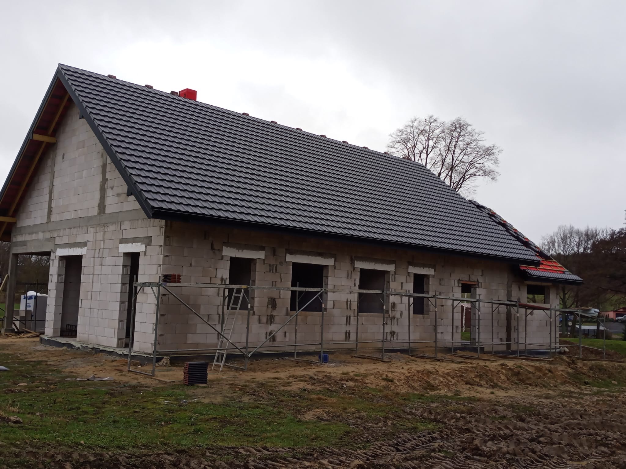Surowy dom z szarej cegły z czarnym dachem z dachówki, otoczony rusztowaniem. Widoczne niezabudowane otwory okienne i drzwiowe. W tle drzewa i pochmurne niebo.