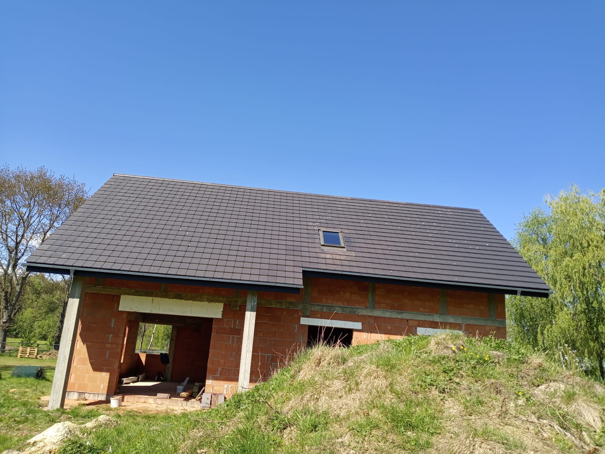 Surowy stan domu z czerwonej cegły z ciemnym dachem, okno dachowe, widok z zewnątrz na tle błękitnego nieba i zieleni. Widać niezadaszone otwory okienne i drzwiowe.